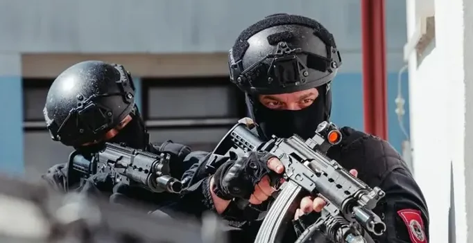 Dva specijalca u crnim uniformama, sa šljemovima i maskama na licu, drže automatske puške u pripravnosti. Fokus je na njihovom naoružanju i opremi, sugerišući operaciju visoke sigurnosti ili obuku.