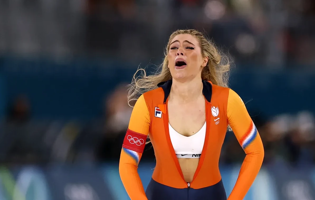 Ženska sportistkinja u narandžastom kombinezonu sa olimpijskim prstenovima na rukavu, sa otvorenim ustima i izrazom iscrpljenosti na licu.