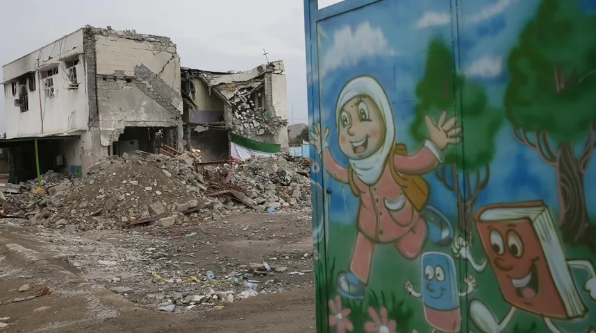 Fotografija prikazuje kontrast između srušene zgrade u pozadini i živopisnog crteža na vratima s nasmijanom djevojčicom u hidžabu, koja nosi ranac, te crtanim likovima knjige i gumice u pokretu.
