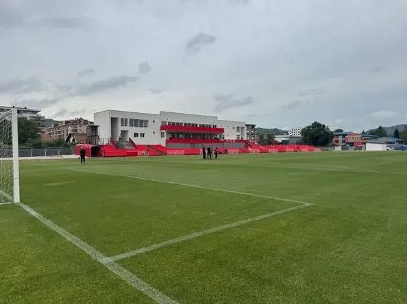 Fudbalski stadion sa novim, zelenim terenom i modernim tribinama obojenim u crveno i bijelo.