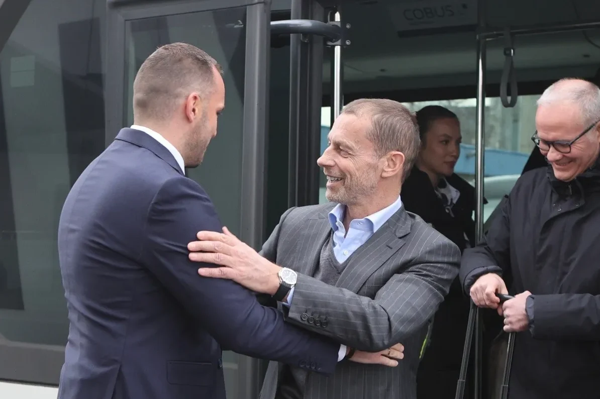 Dvojica muškaraca u odijelima razgovaraju ispred otvorenih vrata autobusa, dok treći muškarac stoji iza njih držeći kofer.