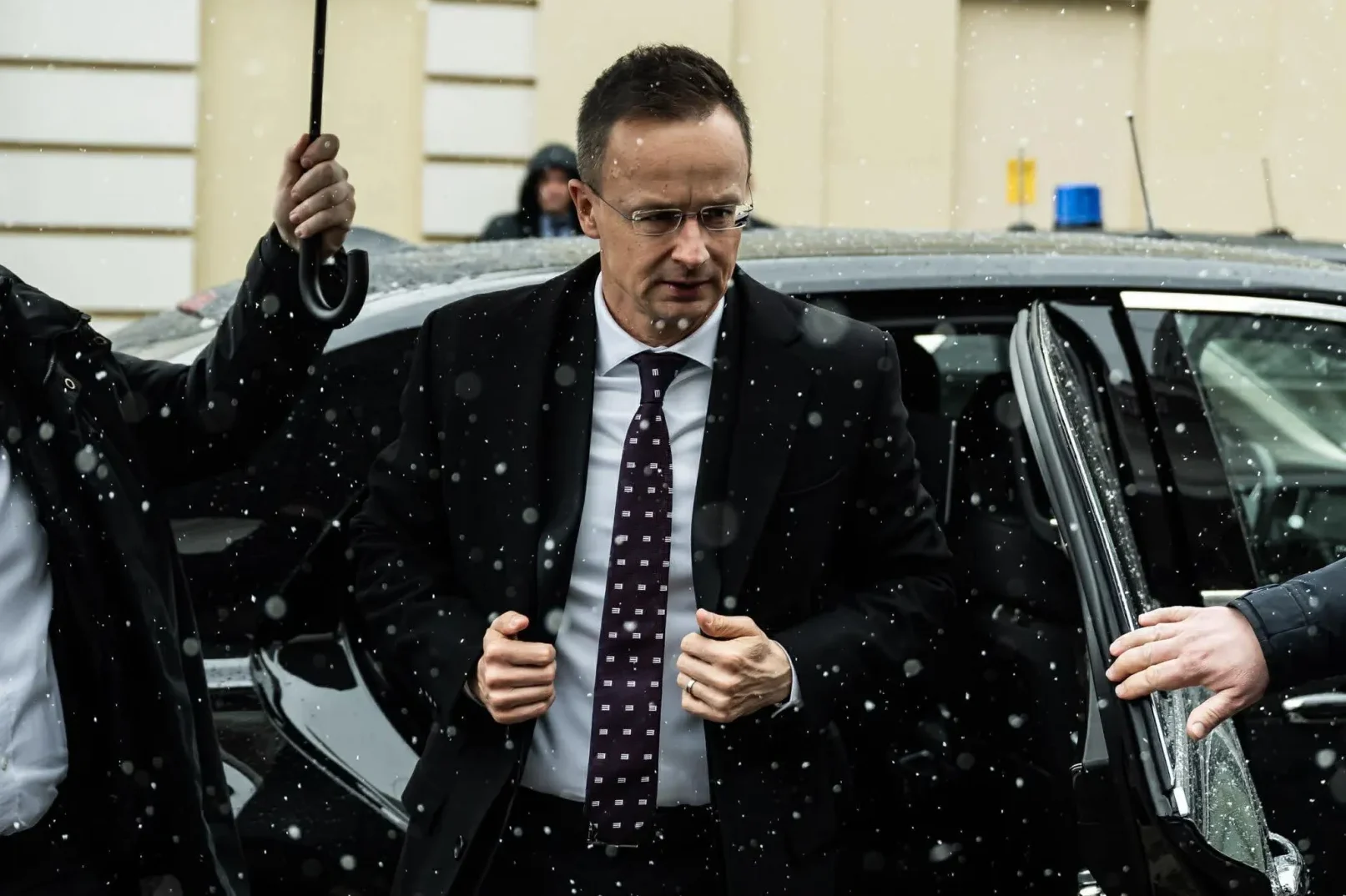 Muškarac u crnom odijelu i bijeloj košulji, sa ljubičastom kravatom, izlazi iz crnog automobila dok pada snijeg. Čini se da su mu pomagači blizu, jedan drži kišobran iznad njega, a drugi otvara vrata automobila.