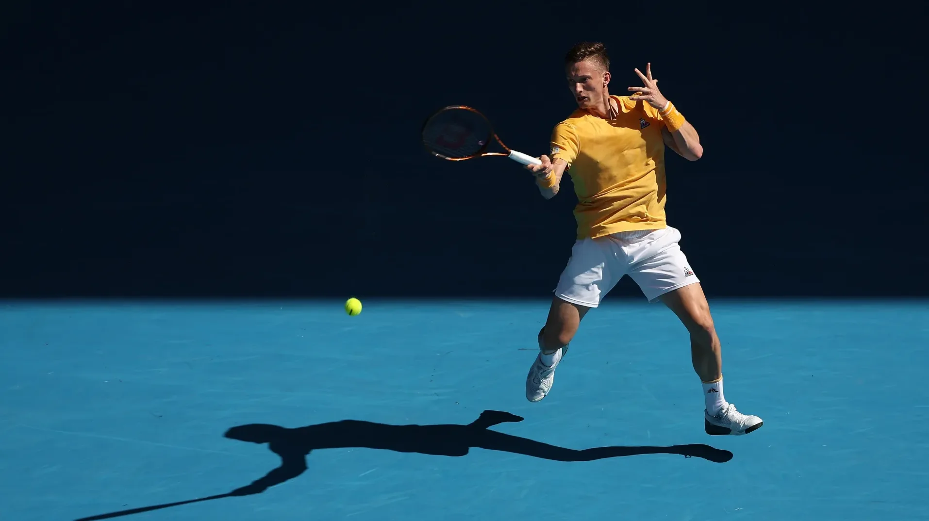 Teniser u žutom dresu i bijelim šortsu zamahuje reketom prema loptici na plavoj teniskoj podlozi.