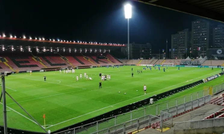 Fudbalski teren osvijetljen reflektorima, igrači u bijelim i plavim dresovima izvode trening.