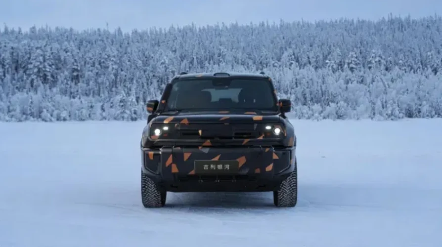 Terensko vozilo sa kamuflažnim dizajnom u narandžastim i crnim tonovima, parkirano na snijegom prekrivenoj ravnici ispred šume prekrivene snijegom.
