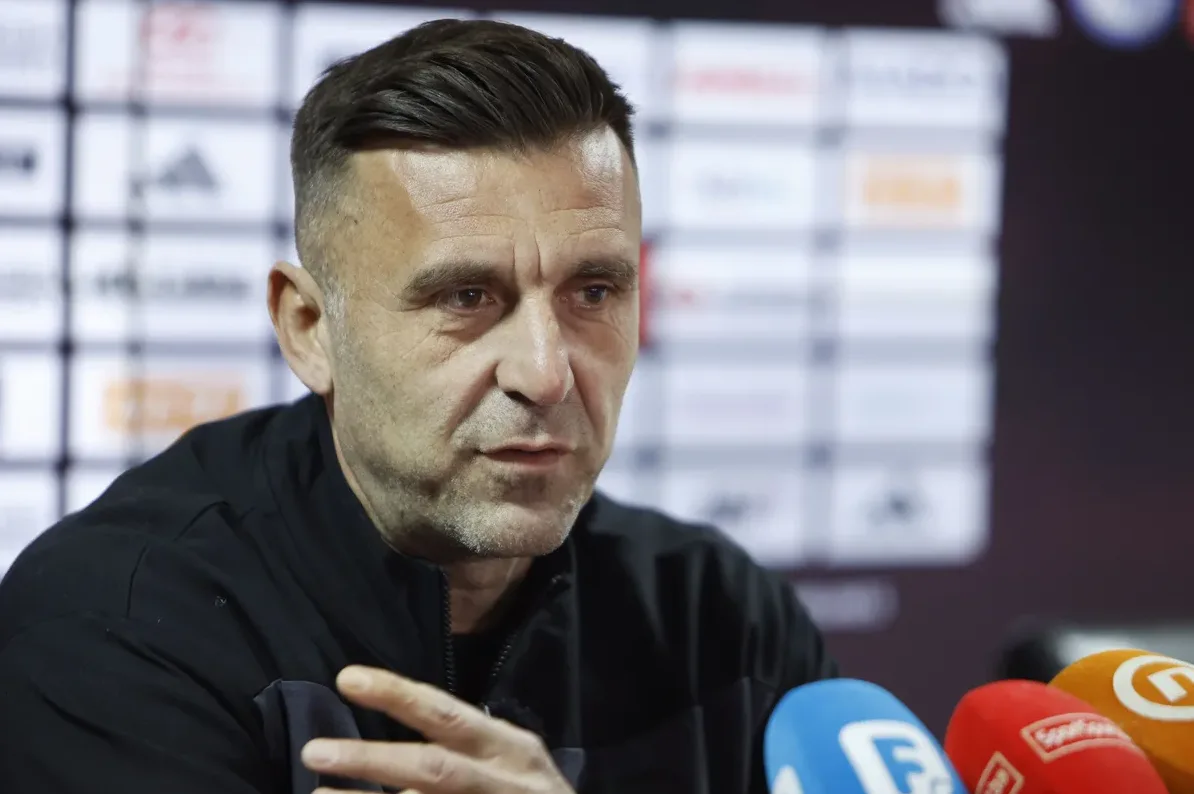 Fudbalski trener sa kratkom kosom, u crnoj jakni, govori na konferenciji za medije ispred mikrofona.