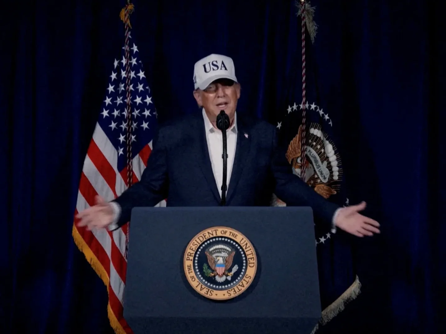 Donald Trump nosi bijelu kapu "USA" i tamni sako, govori sa govornice sa pečatom Predsjednika SAD-a.