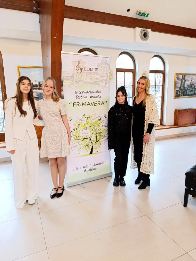 Četiri mlade učesnice festivala "Primavera" stoje ispred banera sa logom UGMUS-a i natpisom "Internacionalni festival muzike "Primavera"" i informacijama o etno selu "Stanišići" Bijeljina.