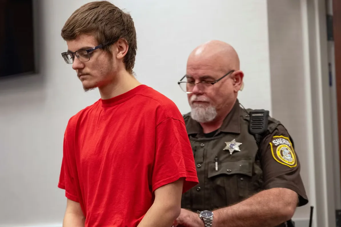 Mladić u crvenoj majici sa naočarima gleda u pod, dok ga prati šerif u uniformi sa oznakama.