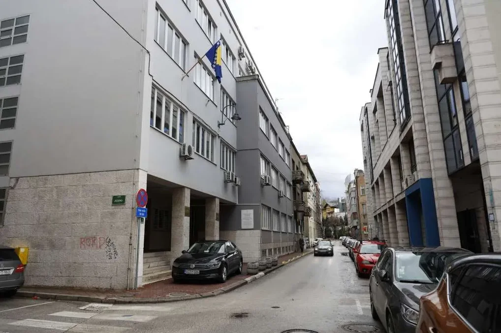 Zgrada sa zastavom Bosne i Hercegovine i parkirani automobili niz ulicu.