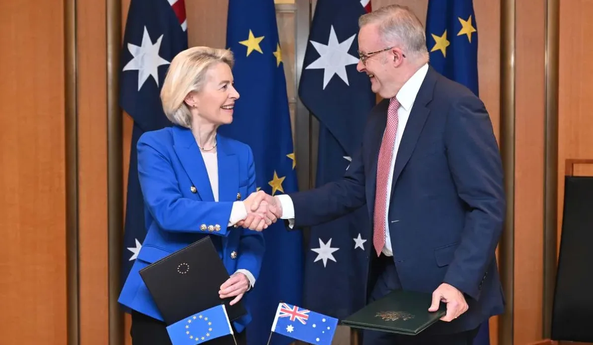 Ursula von der Leyen i Anthony Albanese se rukuju ispred zastava Evropske unije i Australije, oboje drže dokumenta.