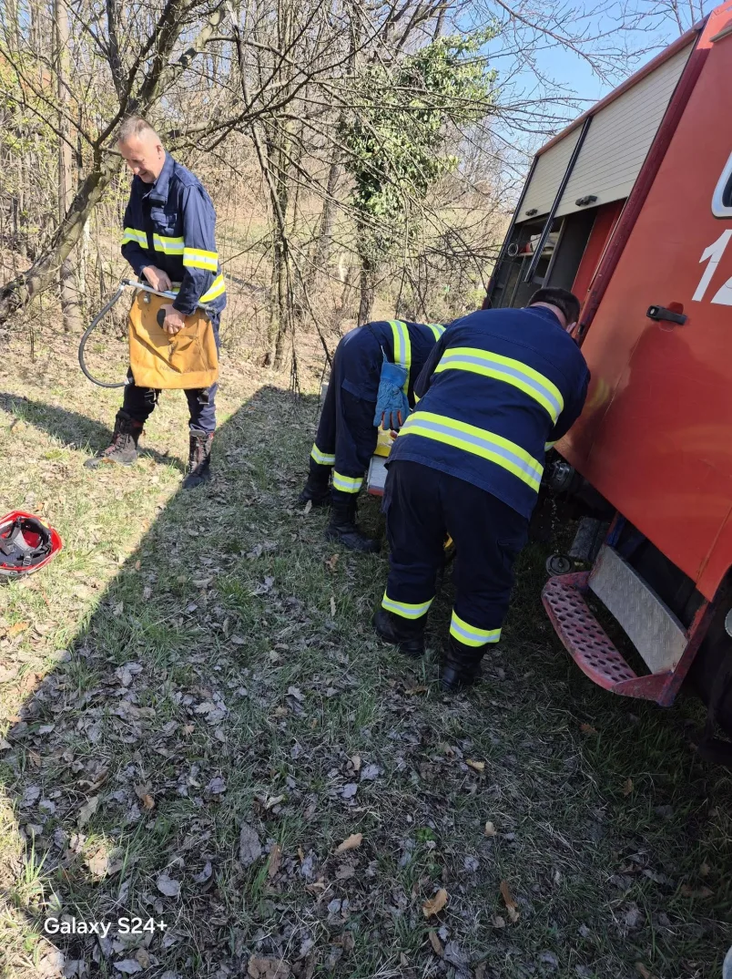 Vatrogasci u radnoj uniformi sa reflektirajućim trakama pregledaju vatrogasno vozilo parkirano na travnatoj površini okruženoj drvećem.