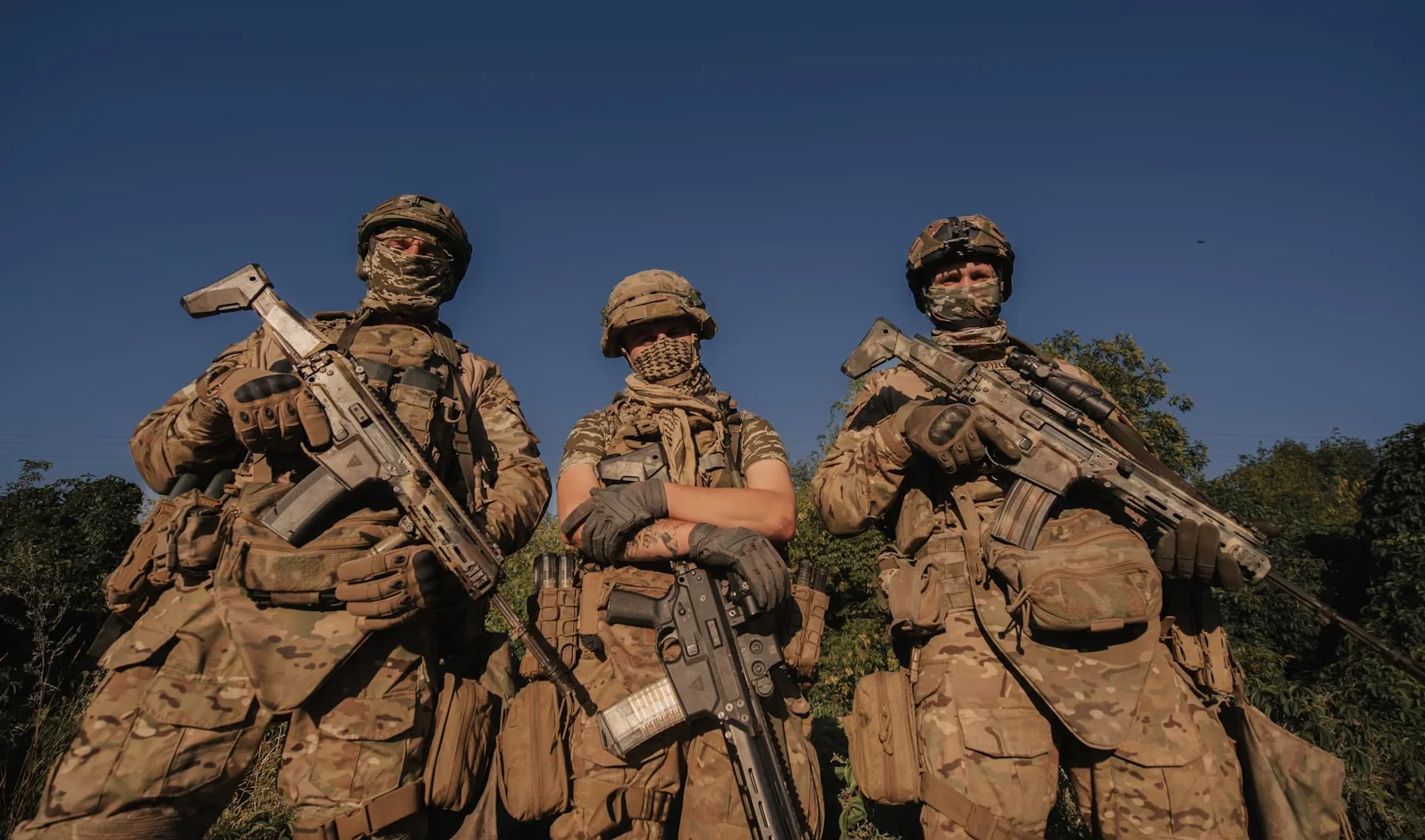 Tri vojnika u maskirnim uniformama i kacigama, naoružani puškama, stoje ispred plavog neba i zelenila.