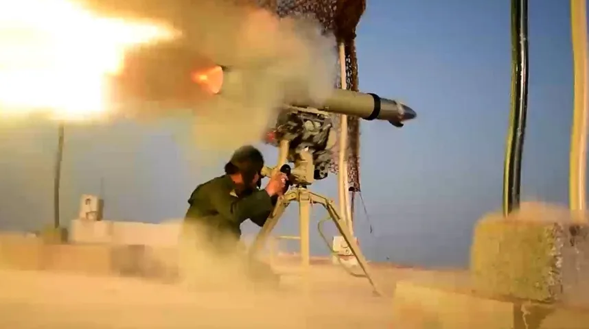 Vojnik u zelenoj uniformi nišani i ispaljuje raketu sa tripod montaže u sumrak, sa plamenom i dimom koji izlaze iz rakete.