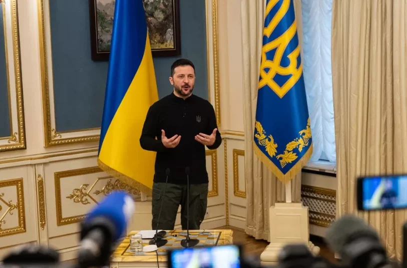 Volodymyr Zelensky, the President of Ukraine, stands and speaks in front of two Ukrainian flags and curtains.