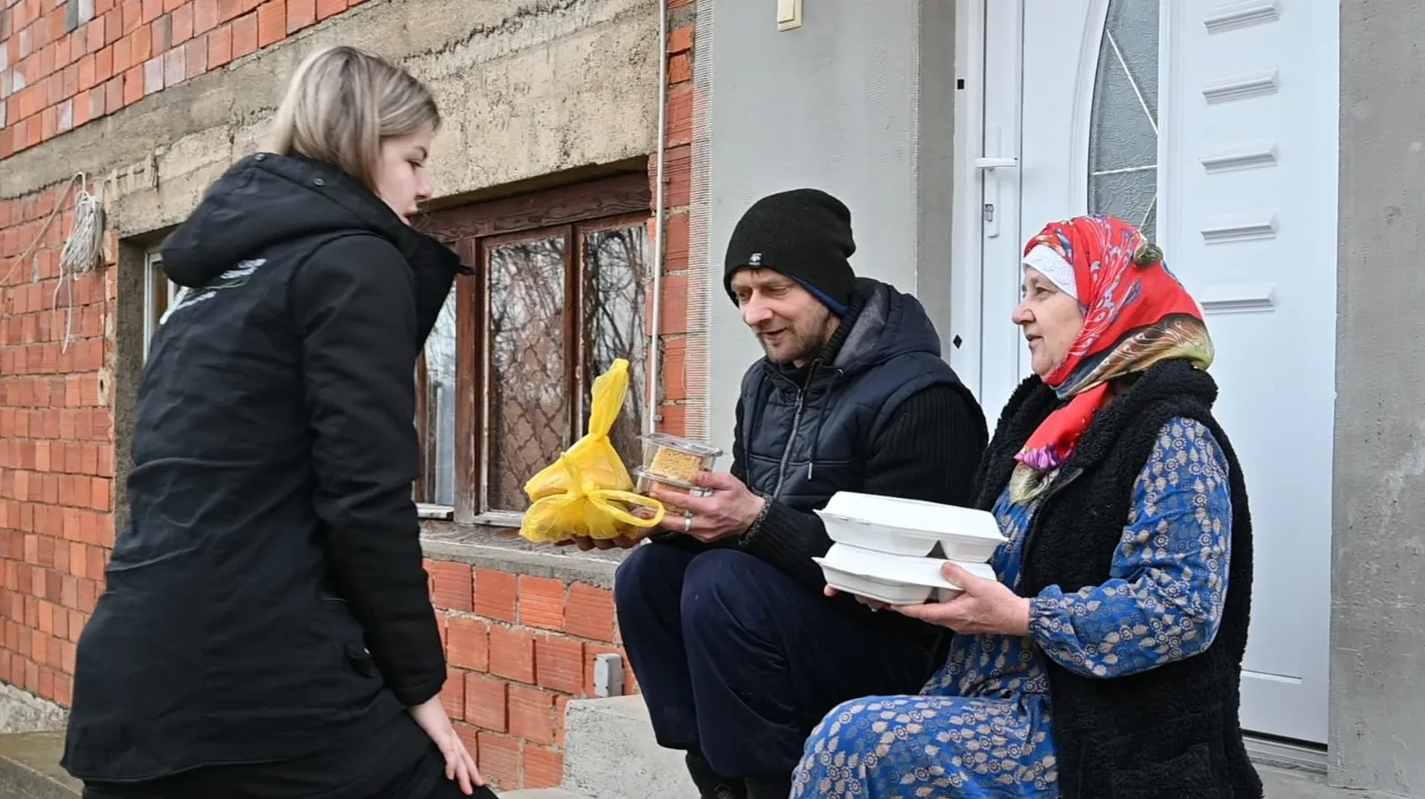 Mlada volonterka u crnoj jakni predaje pakete hrane starijoj ženi sa crvenom maramom na glavi i muškarcu koji sjedi pored nje.