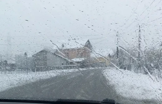 Pogled kroz šoferšajbu automobila prekrivenu kapljicama vode i snijegom, sa zgradama i drvećem u pozadini.