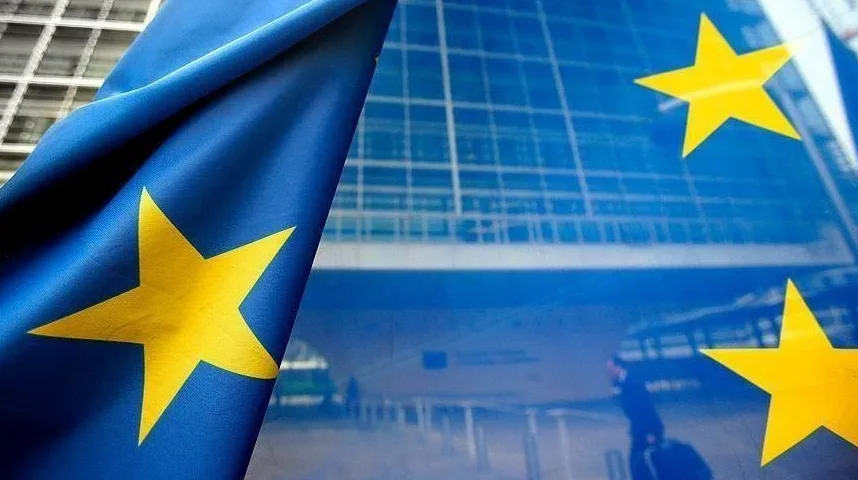 Zastava Evropske unije sa zlatnim zvijezdama na plavoj podlozi, postavljena ispred moderne staklene zgrade institucija EU.