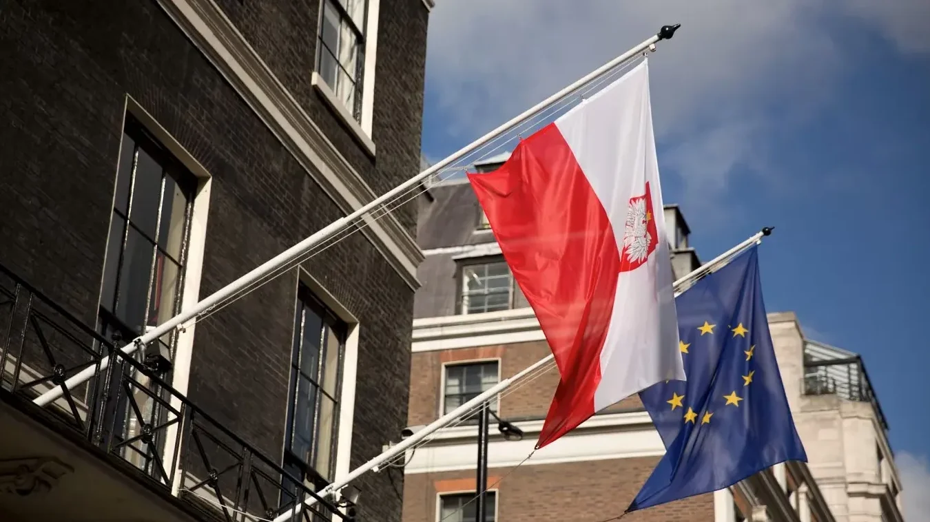 Zastava Poljske i zastava Evropske Unije istaknute na jarbolima ispred tamne ciglene zgrade.