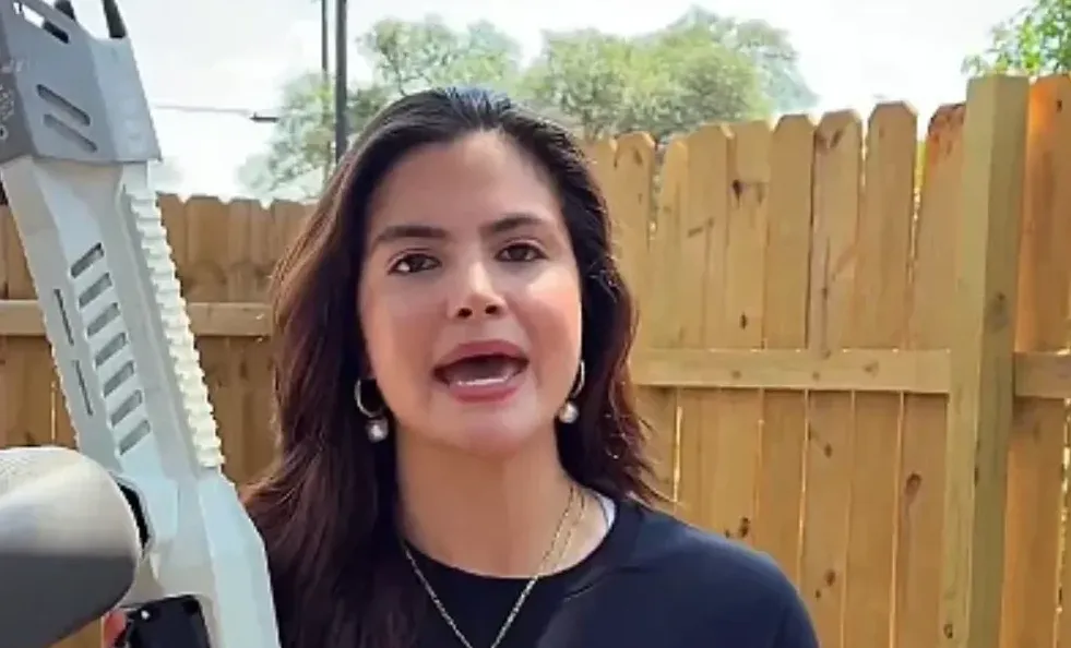 Žena drži bijelu airsoft ili paintball pušku dok govori, a u pozadini je drvena ograda i drveće.