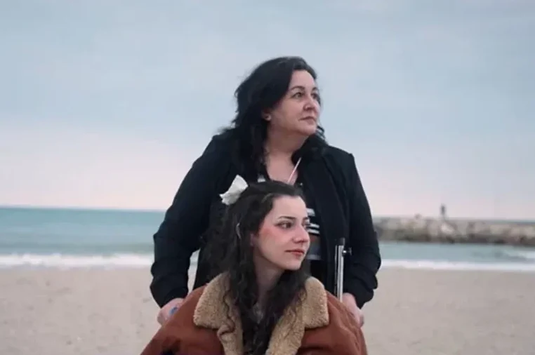 Žena gura djevojku u invalidskim kolicima na pješčanoj plaži pored mora.