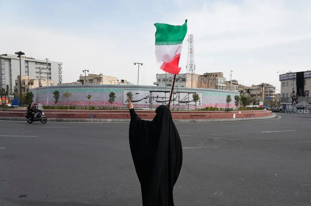 Žena u crnom hidžabu drži iransku zastavu podignutu u zrak na raskrsnici.