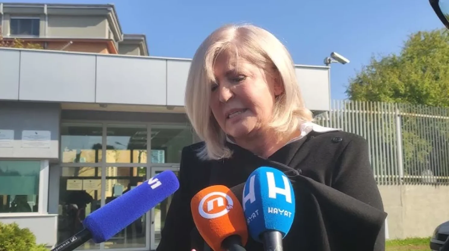 Žena sa plavom kosom govori u mikrofone ispred zgrade.