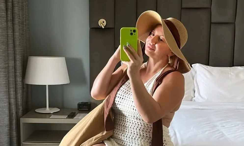 Žena u ljetnom šeširu i bijeloj kukičanoj haljini slika se telefonom u hotelskoj sobi.