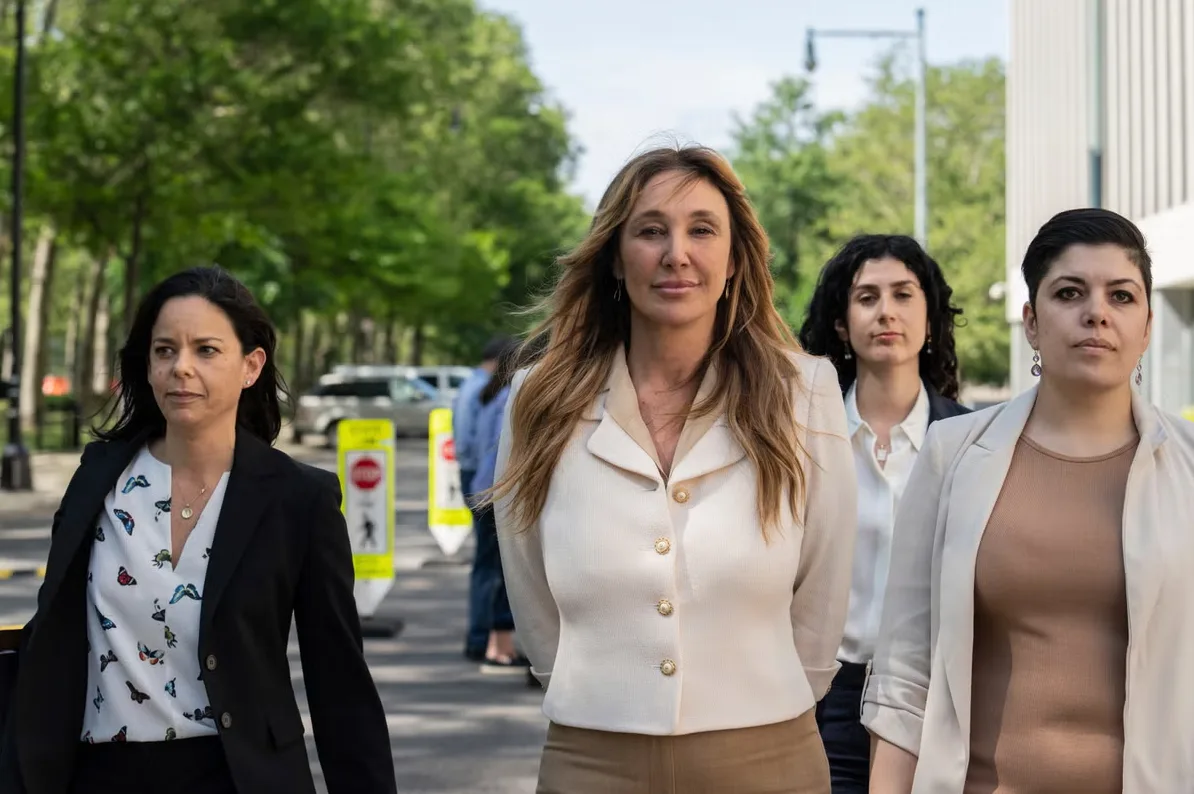 Četiri žene u elegantnoj odjeći izlaze iz zgrade, predvođene ženom sa dugom plavom kosom i blijedo bež sakoom.