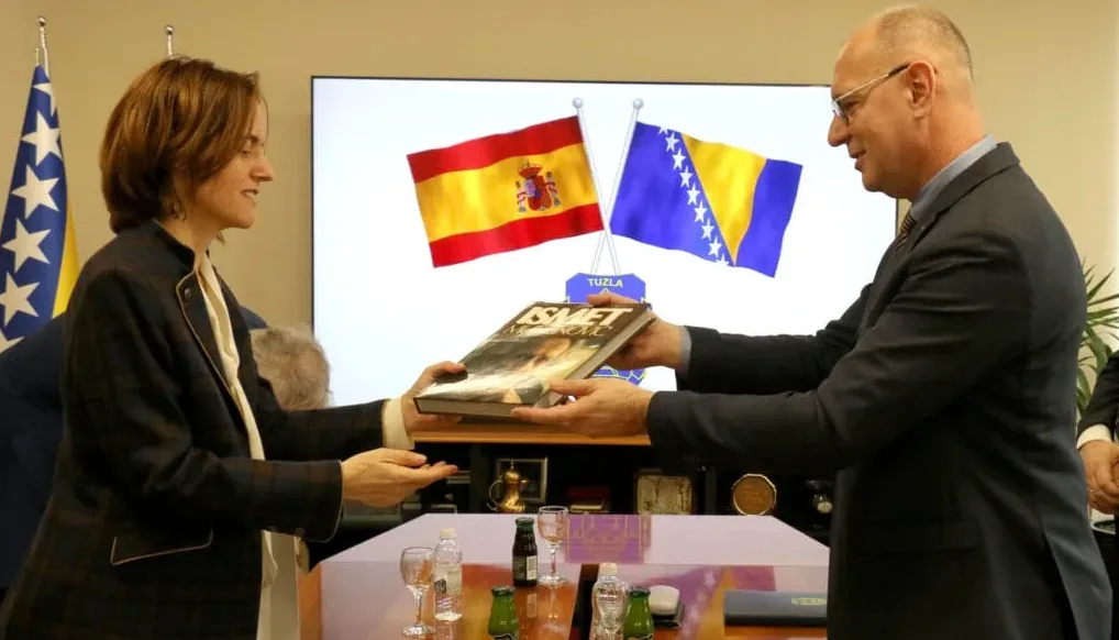 Dvije osobe razmjenjuju knjigu ispred zastava Španije i Bosne i Hercegovine.
