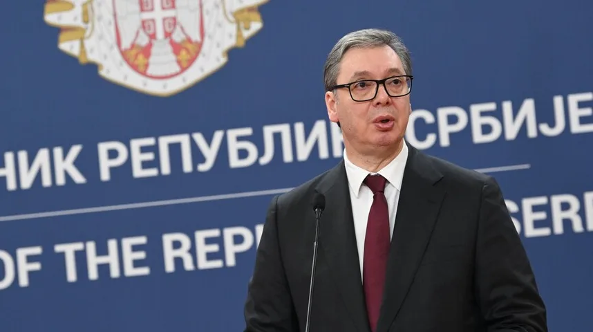 Aleksandar Vučić nosi tamno odijelo i crvenu kravatu, govoreći za govornicom ispred grba Republike Srbije.