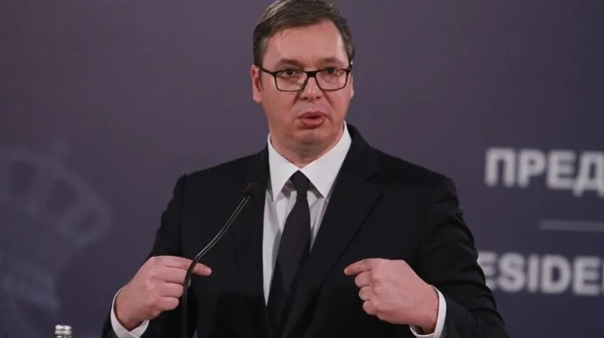Aleksandar Vučić u crnom odijelu i bijeloj košulji sa crnom kravatom, sa naočarima, govori ispred mikrofona, pokazujući prstima na sebe. U pozadini je tamna pozadina sa natpisom na ćirilici.