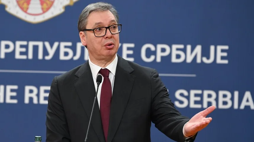 Aleksandar Vučić u crnom odijelu i tamno crvenoj kravati, s naočarima, govori za govornicom ispred plave pozadine sa natpisom "REPUBLIKA SRBIJE".