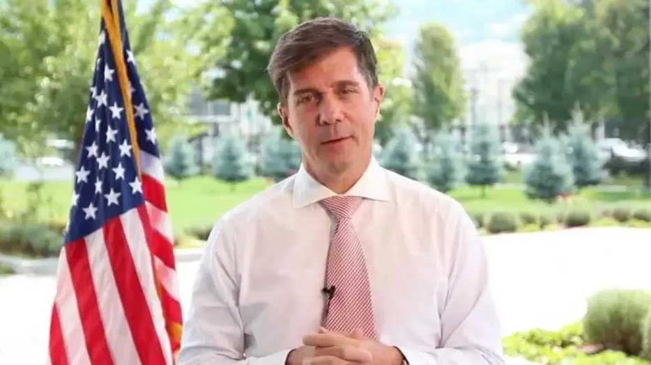 Političar ili javna ličnost, obučen u bijelu košulju i crvenu prugastu kravatu, stoji ispred američke zastave i zelenila u pozadini.