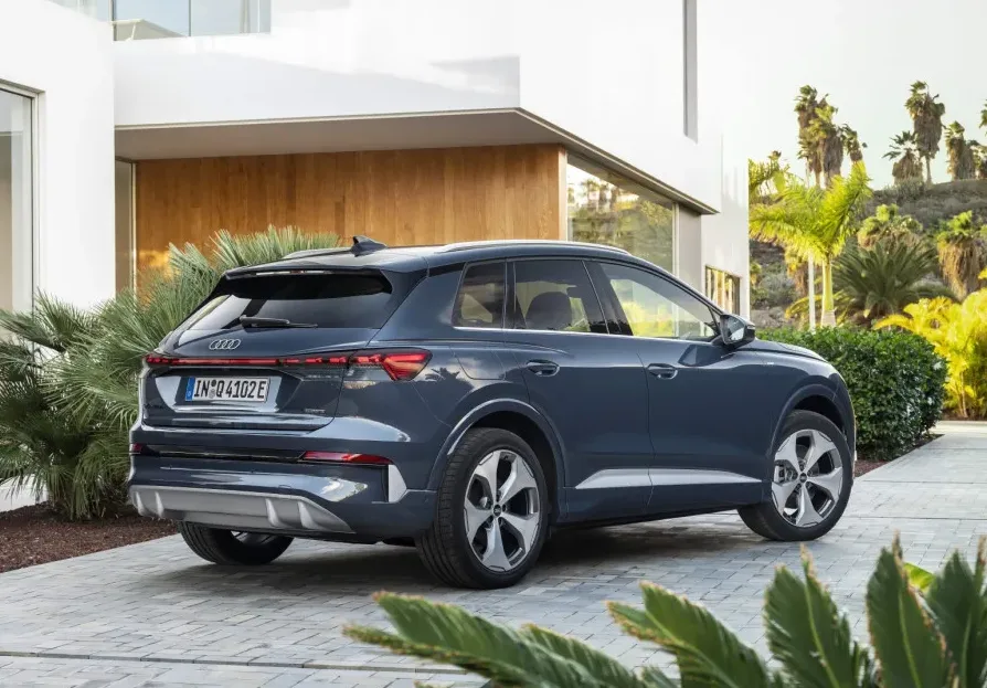 Tamno plavi Audi Q4 e-tron SUV iz 2023. godine parkiran ispred moderne bijele zgrade sa bujnim zelenilom i palmama u pozadini.