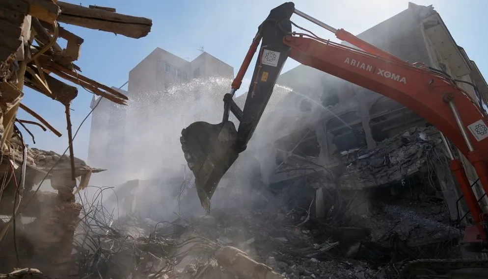 Bager marke Arian Xoma sa kašikom podignutom u vazduh, vrši rušenje oštećene zgrade uz dosta prašine i raspršene vode.