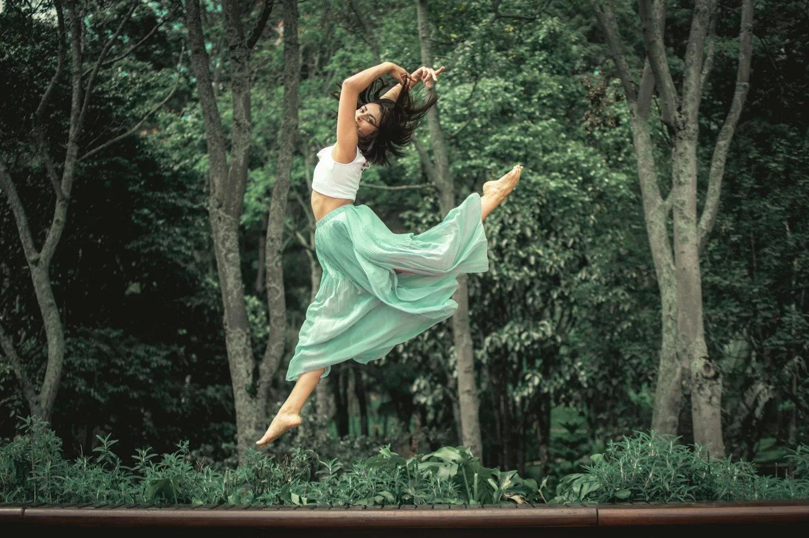 Žena izvodi plesni skok u prostranoj zelenoj suknji i bijelom topu, sa rukama podignutim iznad glave i nogama savijenim u zraku.
