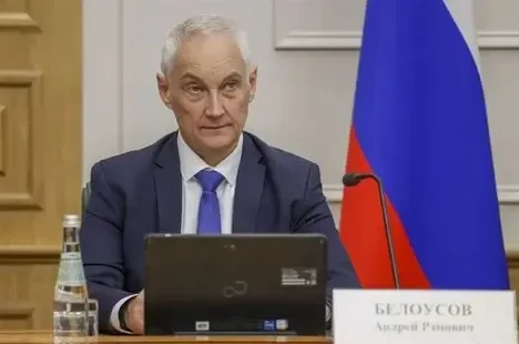 Portret Andreja Belousova, čovjeka sijede kose, u tamnoplavom odijelu i plavoj kravati, kako sjedi za stolom ispred ruske zastave.