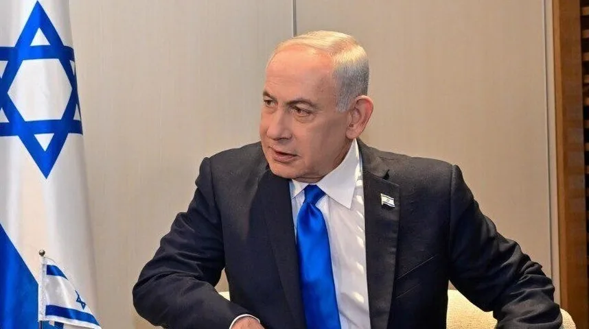 Benjamin Netanyahu u crnom odijelu sa plavom kravatom, sjedi pored zastave Izraela.