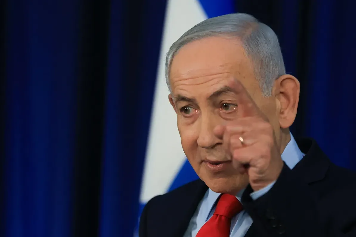Benjamin Netanyahu u tamnom odijelu i crvenoj kravati, pokazuje kažiprstom desne ruke prema naprijed, sa izraelskom zastavom u pozadini.