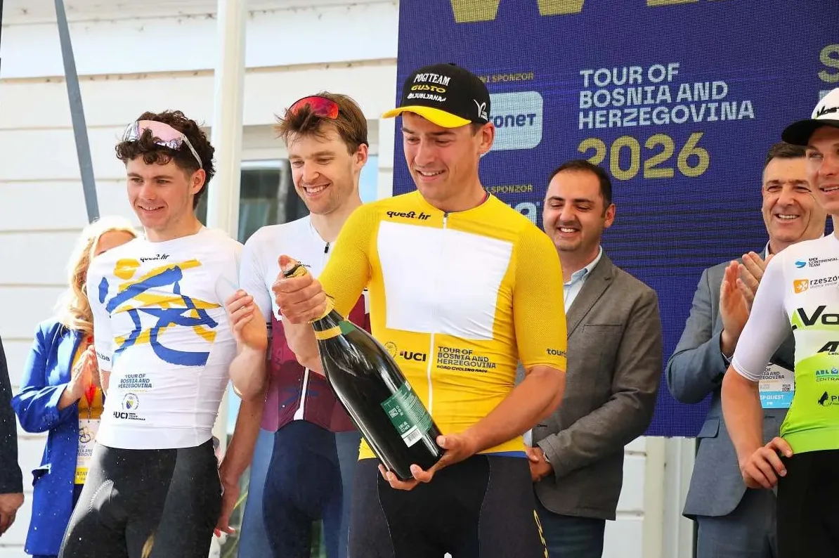 Biciklista u žutom dresu drži veliku bocu pjenušavog vina na podijumu, s osmijehom na licu, na dodjeli nagrada biciklističke utrke "Tour of Bosnia and Herzegovina 2026".