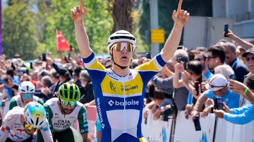 Biciklista u plavo-žutom dresu sa podignutim rukama u znak pobjede, sa publikom u pozadini.