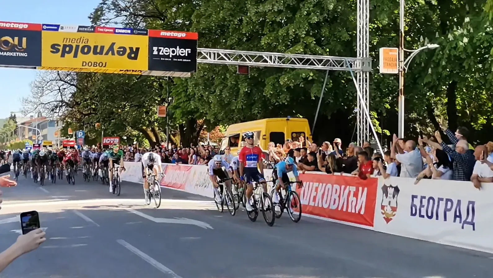 Biciklistička trka sa grupom takmičara kako prelaze ciljnu liniju ispred publike i sponzorskih banera.