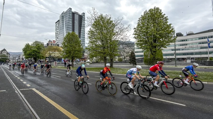 Grupacija biciklista u jarkoj odjeći takmiči se na ulici ispred modernih zgrada.