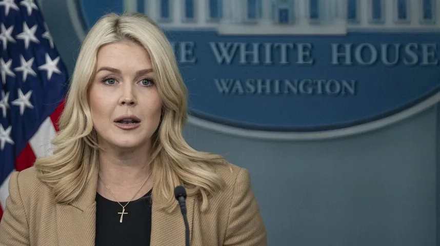 Žena s plavom kosom govori ispred mikrofona, s američkom zastavom i logom Bijele kuće u pozadini.