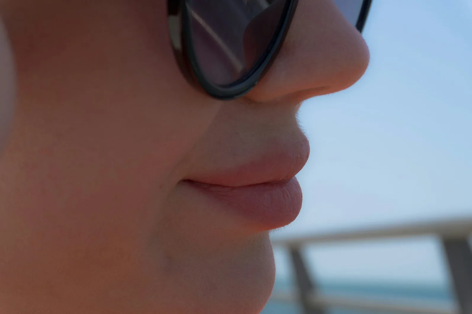 Krupni kadar dijela lica osobe sa sunčanim naočarima i ustima, iz profila, sa plavim nebom u pozadini.