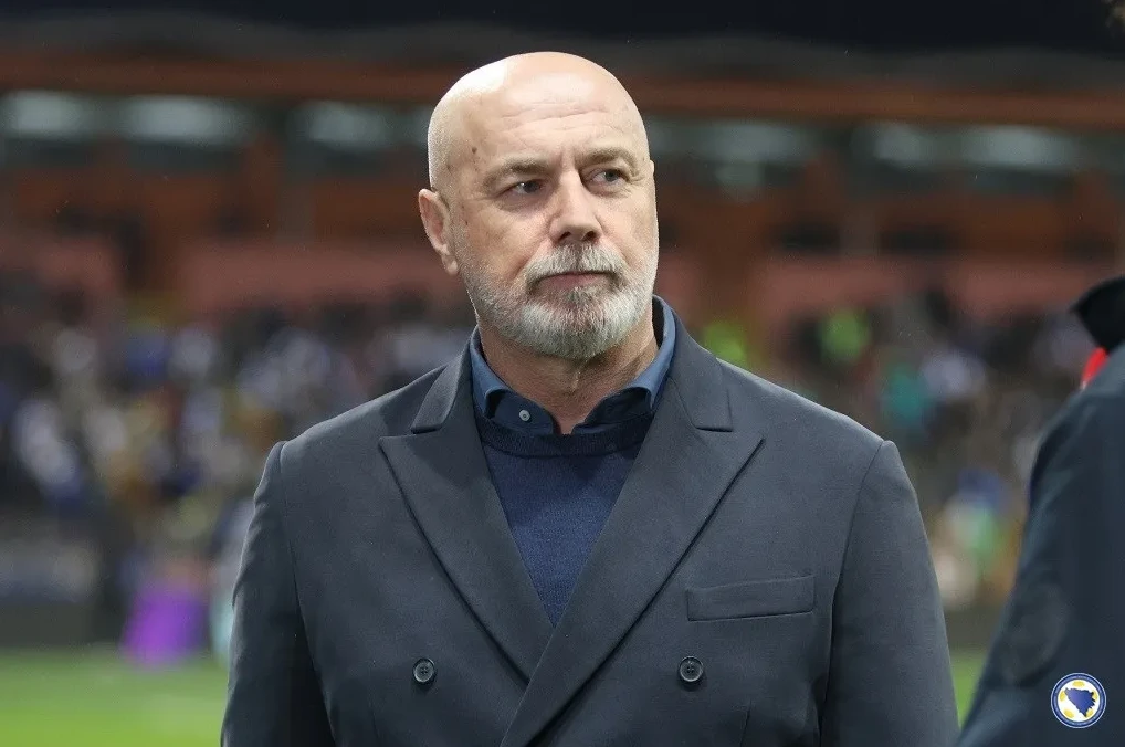 Ćelavi muškarac sa sijedom bradom u tamnom sakou i plavoj rolci, trener fudbalske reprezentacije Bosne i Hercegovine, Ivan Vučević, gleda u daljinu na fudbalskoj utakmici.