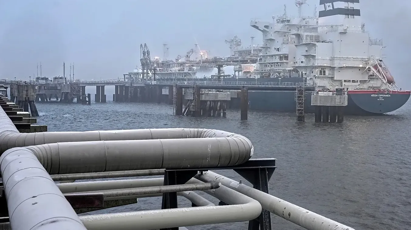 Veliki brod-tanker za tečni gas (LNG) usidren u blizini industrijskog postrojenja sa vidljivim cjevovodima u prvom planu.