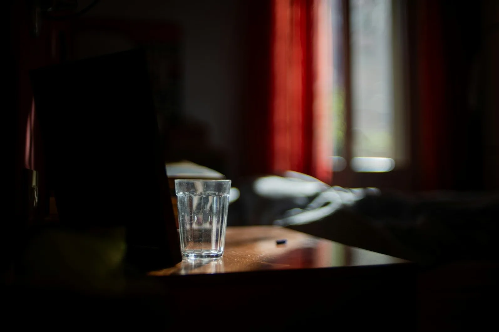 Staklena čaša sa vodom na drvenoj površini osvijetljena sunčevom svjetlošću koja prolazi kroz crveni zastor.
