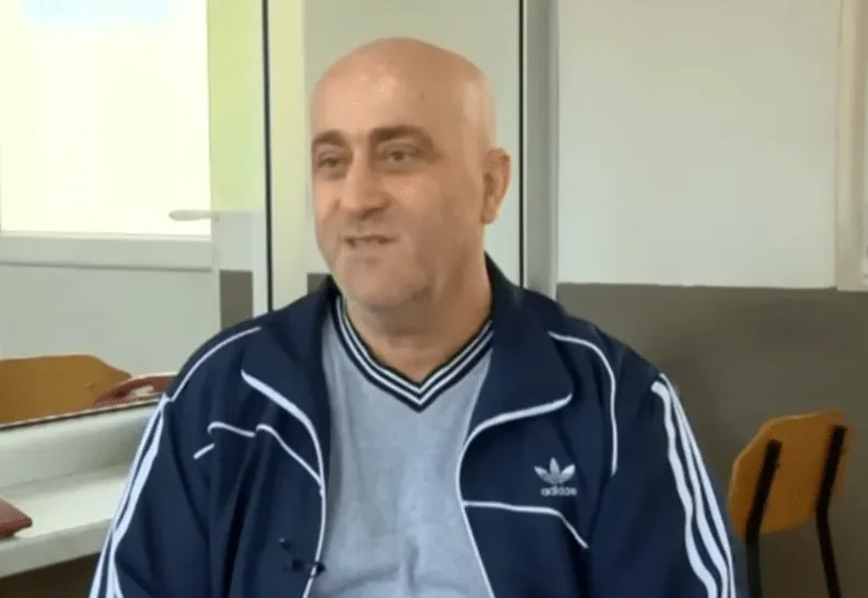 Ćelavi muškarac sa kratkom bradom nosi sivu majicu i tamnoplavu Adidas jaknu sa bijelim prugama na rukavima.